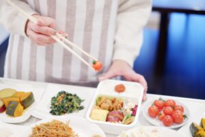 A person picking up a cherry tomato with chopsticks while making a Japanese bento filled with rice, fried chicken, tamagoyaki, macaroni salad and vegetables.