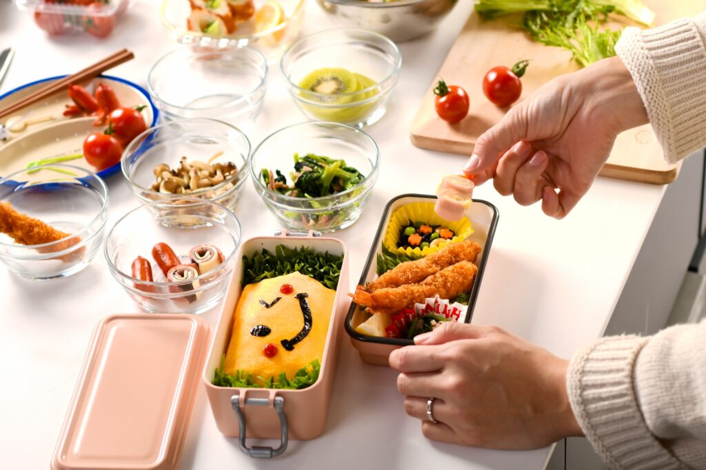 A person assembling a Japanese bento, adding side dishes to a lunchbox containing fried shrimp, vegetables, rolled ham, and a smiling omelet decorated with nori and cherry tomatoes.